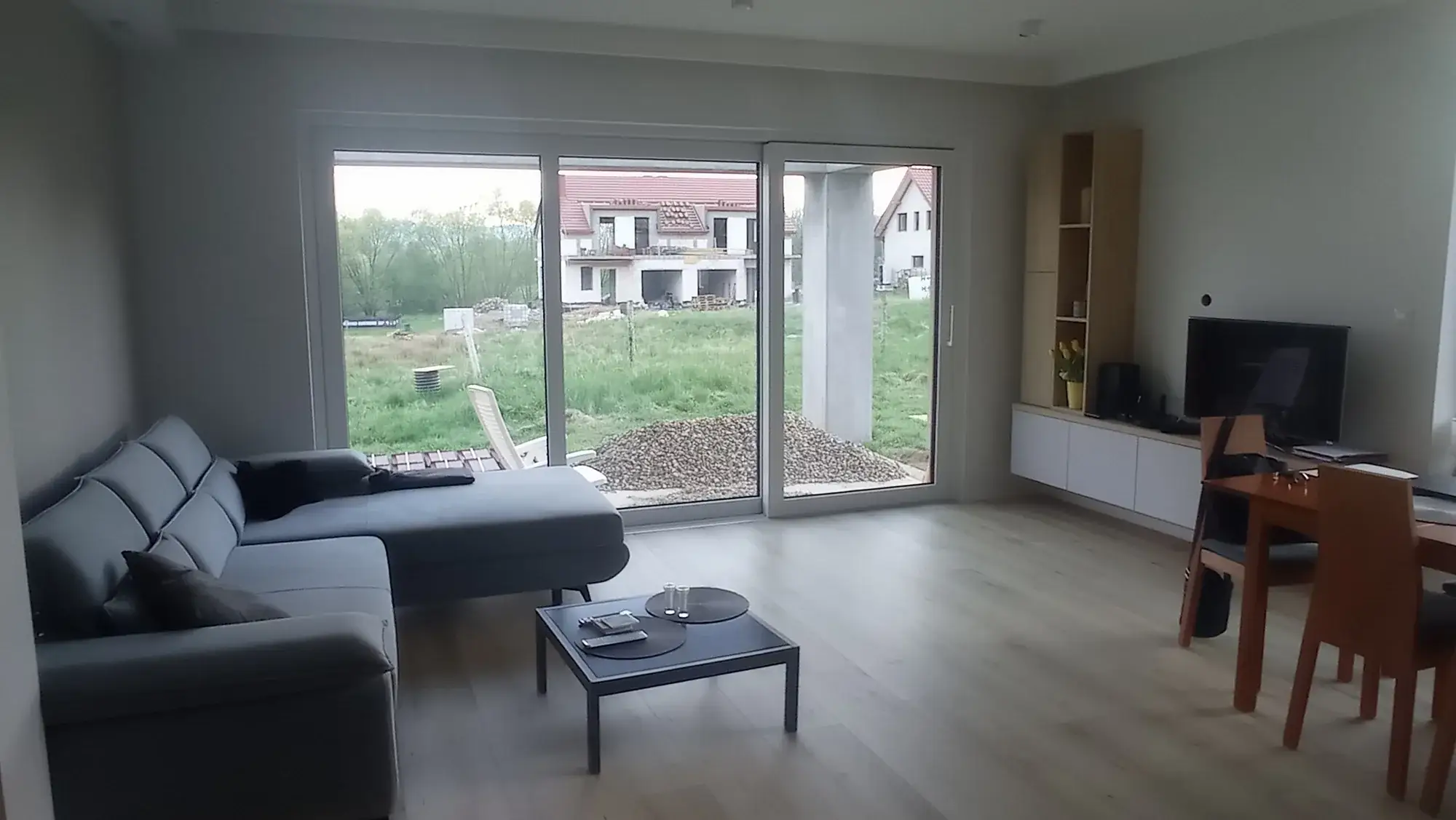 Salon przed metamorfozą: szary narożnik, niski stolik i duże okno na ogród w budowie
