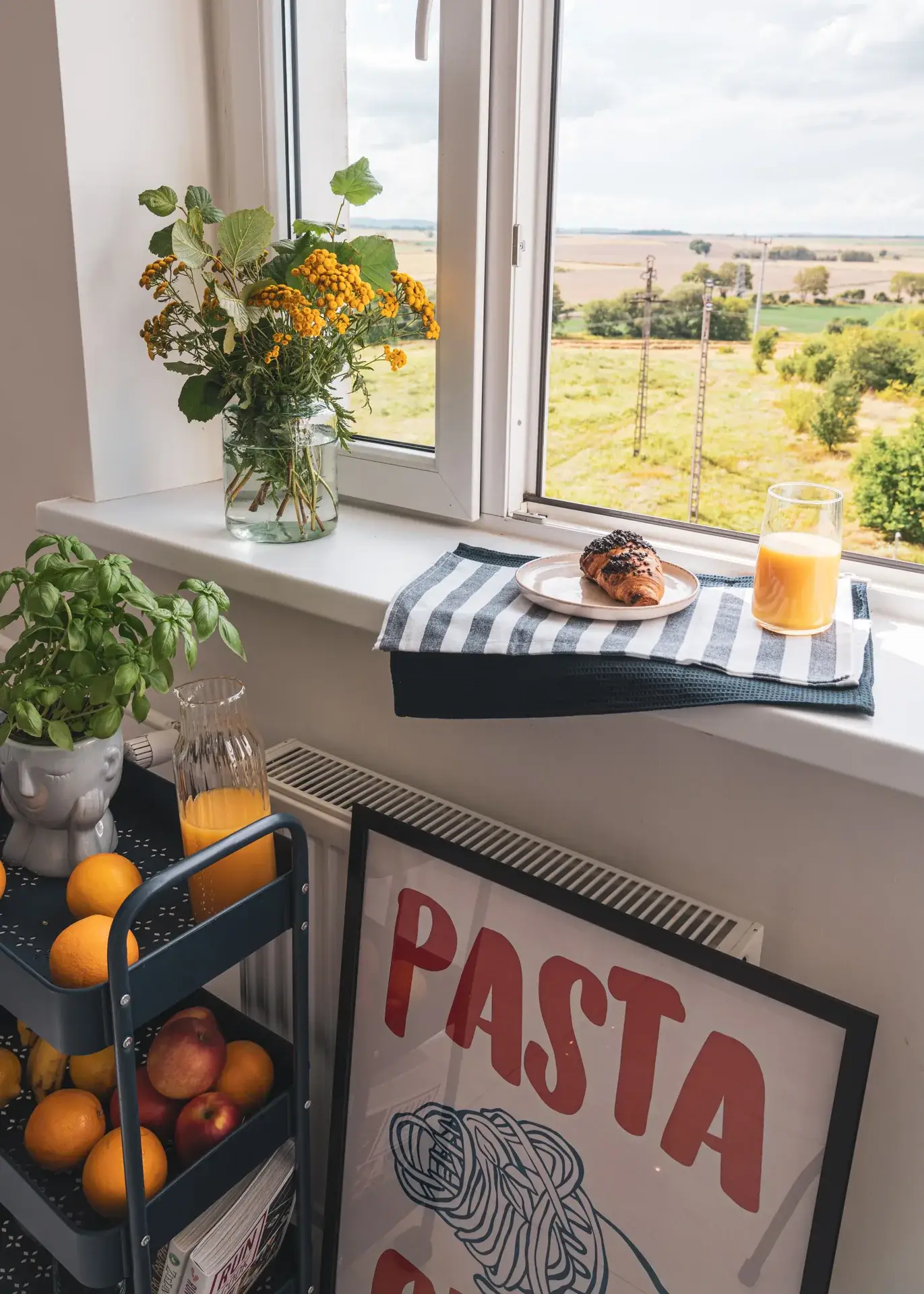Kuchnia po home stagingu: parapet z kwiatami i śniadaniem, widok z okna na zieleń, wózek z owocami i plakat