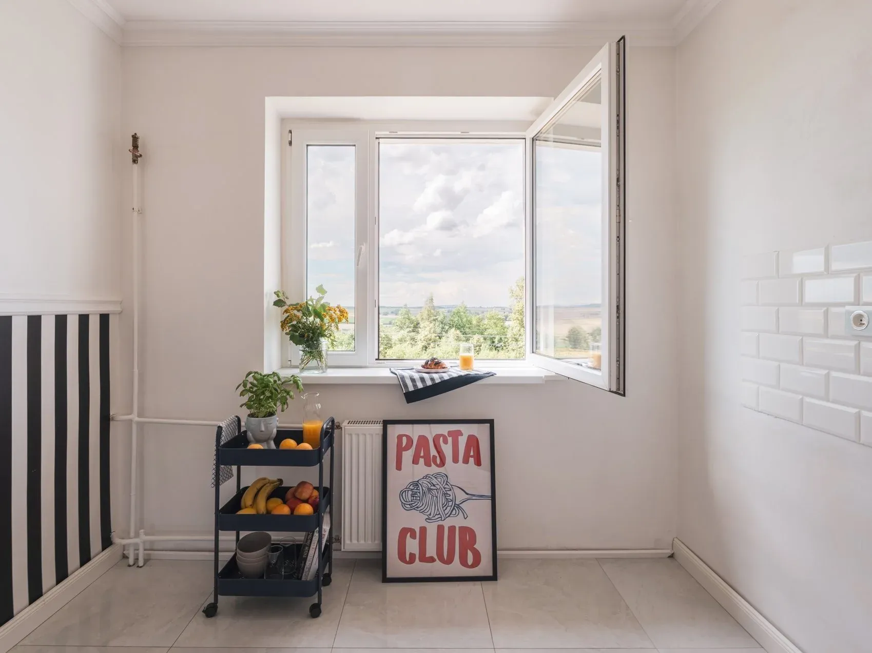 Kuchnia po home stagingu z tego samego miejsca co zdjęcie przed: otwarte okno z widokiem na zieleń, wózek z owocami, plakat Pasta Club i tapeta w pasy