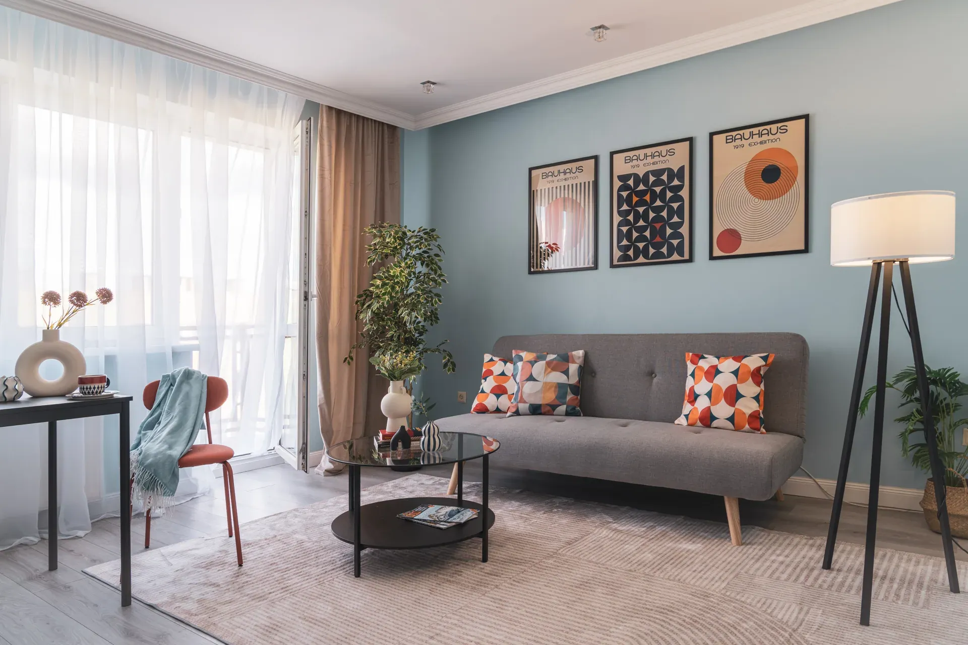 Salon po home stagingu: szara sofa z poduszkami we wzory, szklany stolik, plakaty w stylu Bauhausu i lampa na trójnogu
