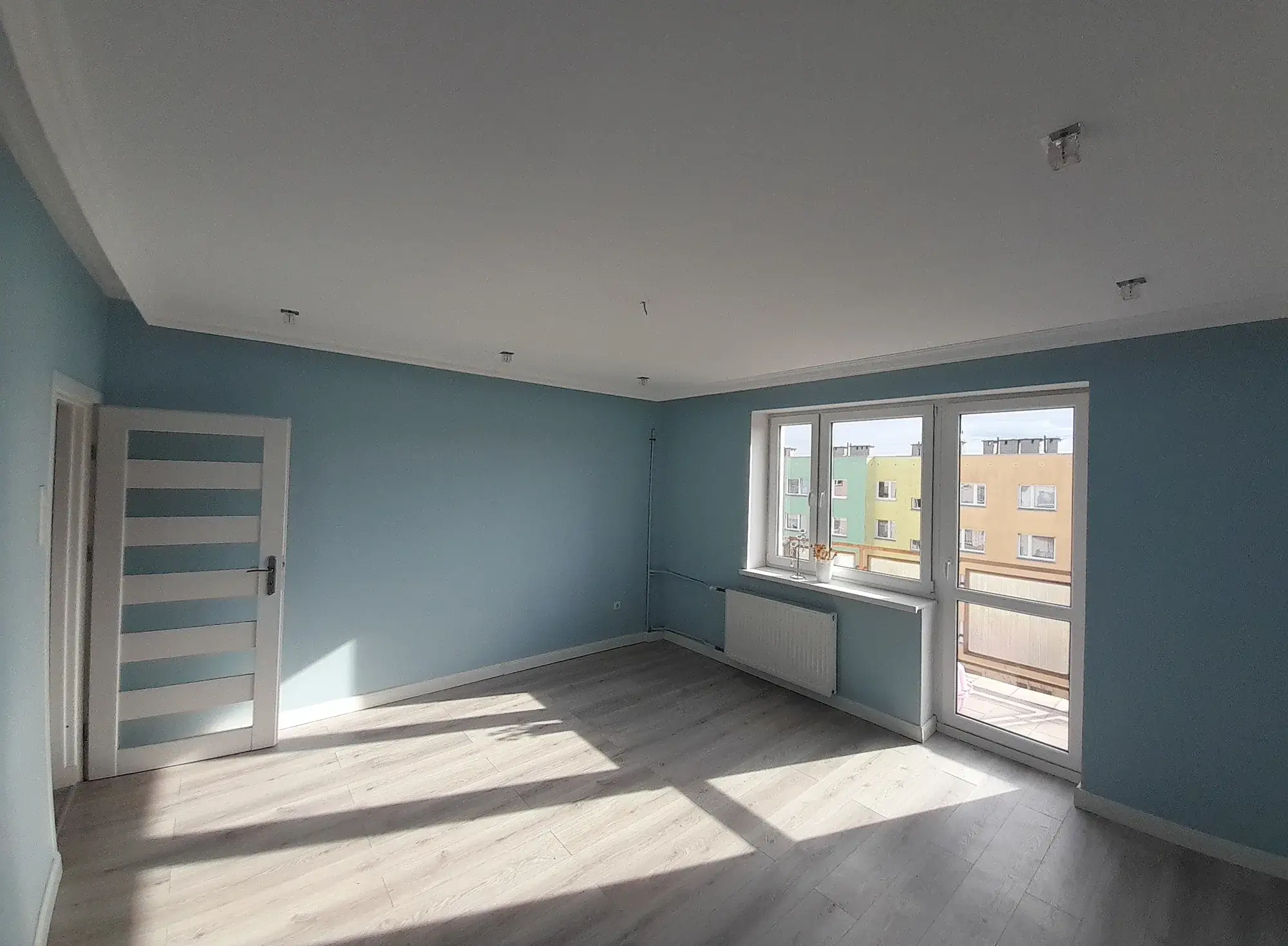 Pusty salon z niebieskimi ścianami przed home stagingiem, widok na okno balkonowe
