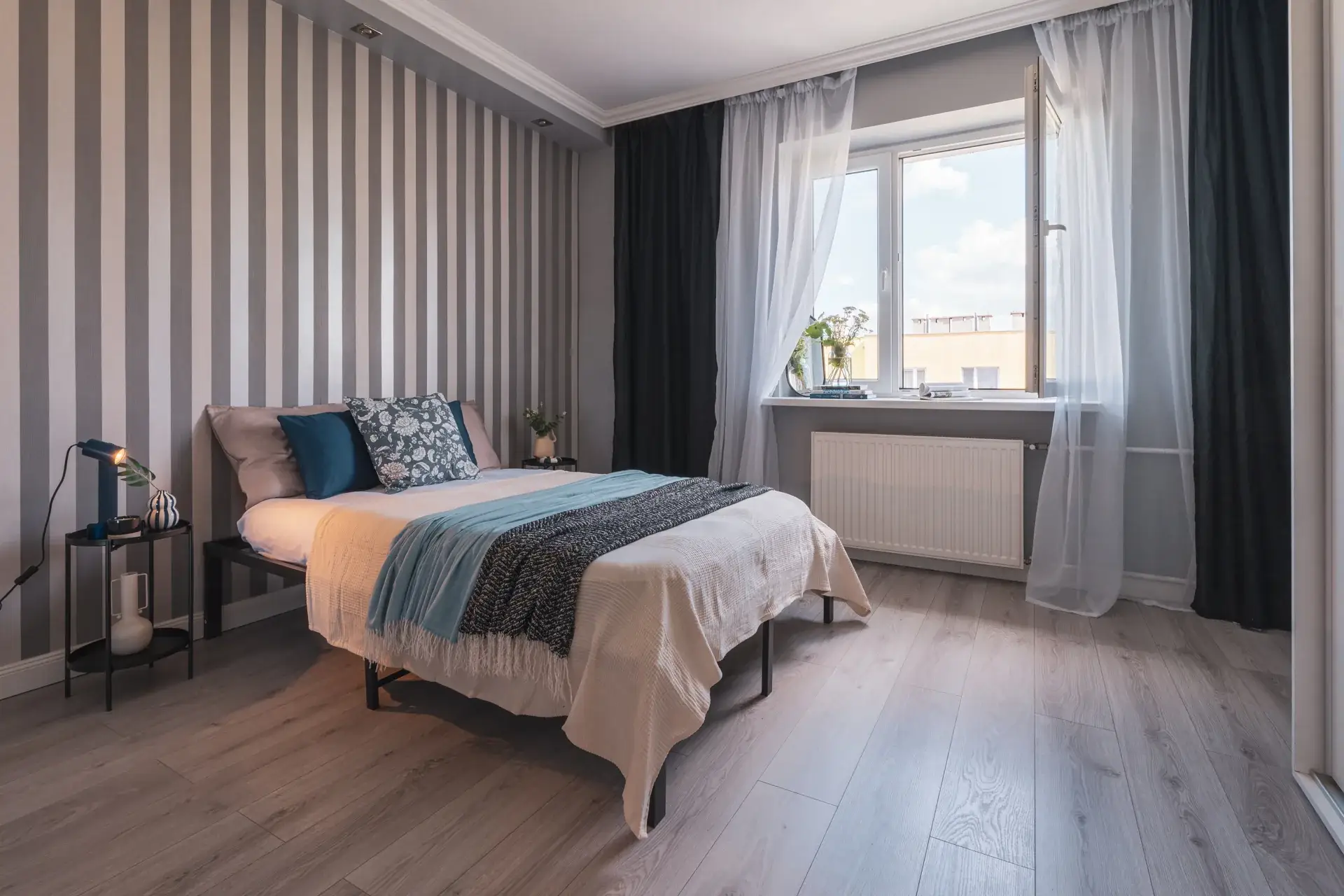 Sypialnia po home stagingu: łóżko z jasną pościelą i niebieskimi poduszkami, rozsunięta firanka, czarne stoliki przy łóżku