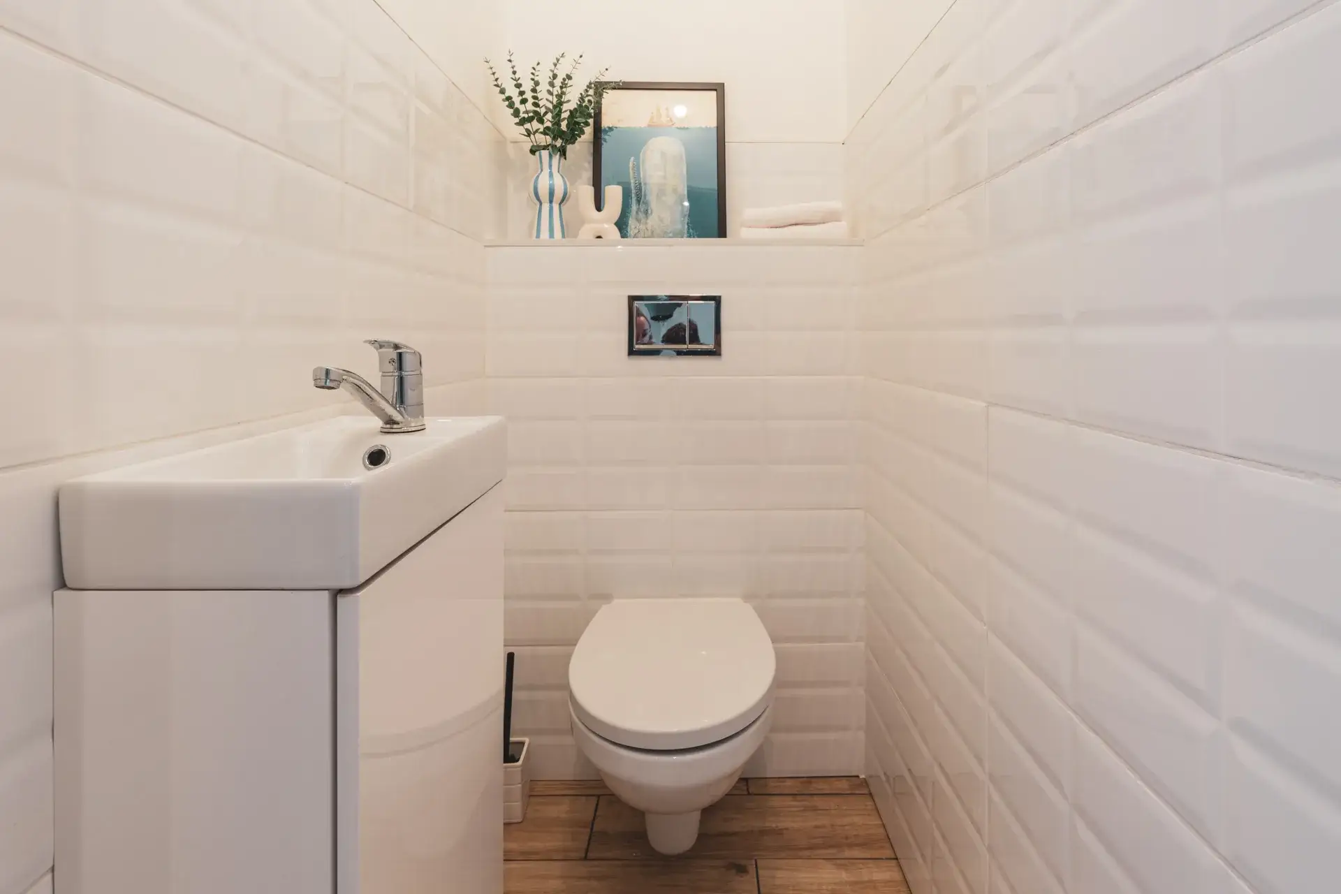 WC po home stagingu: białe kafle, półka z grafiką z wielorybem i gałązką eukaliptusa w wazonie