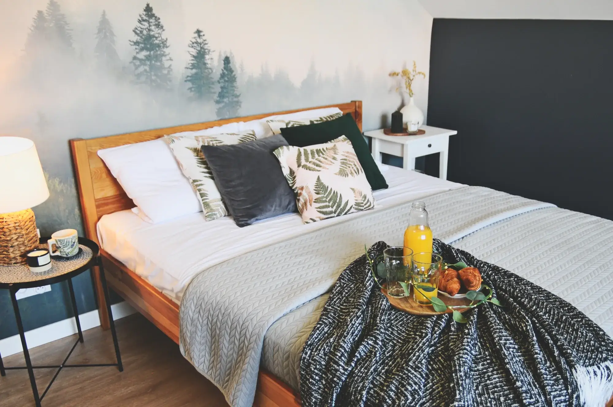 Sypialnia po home stagingu: drewniane łóżko z białą pościelą, taca ze śniadaniem, fototapeta z lasem we mgle