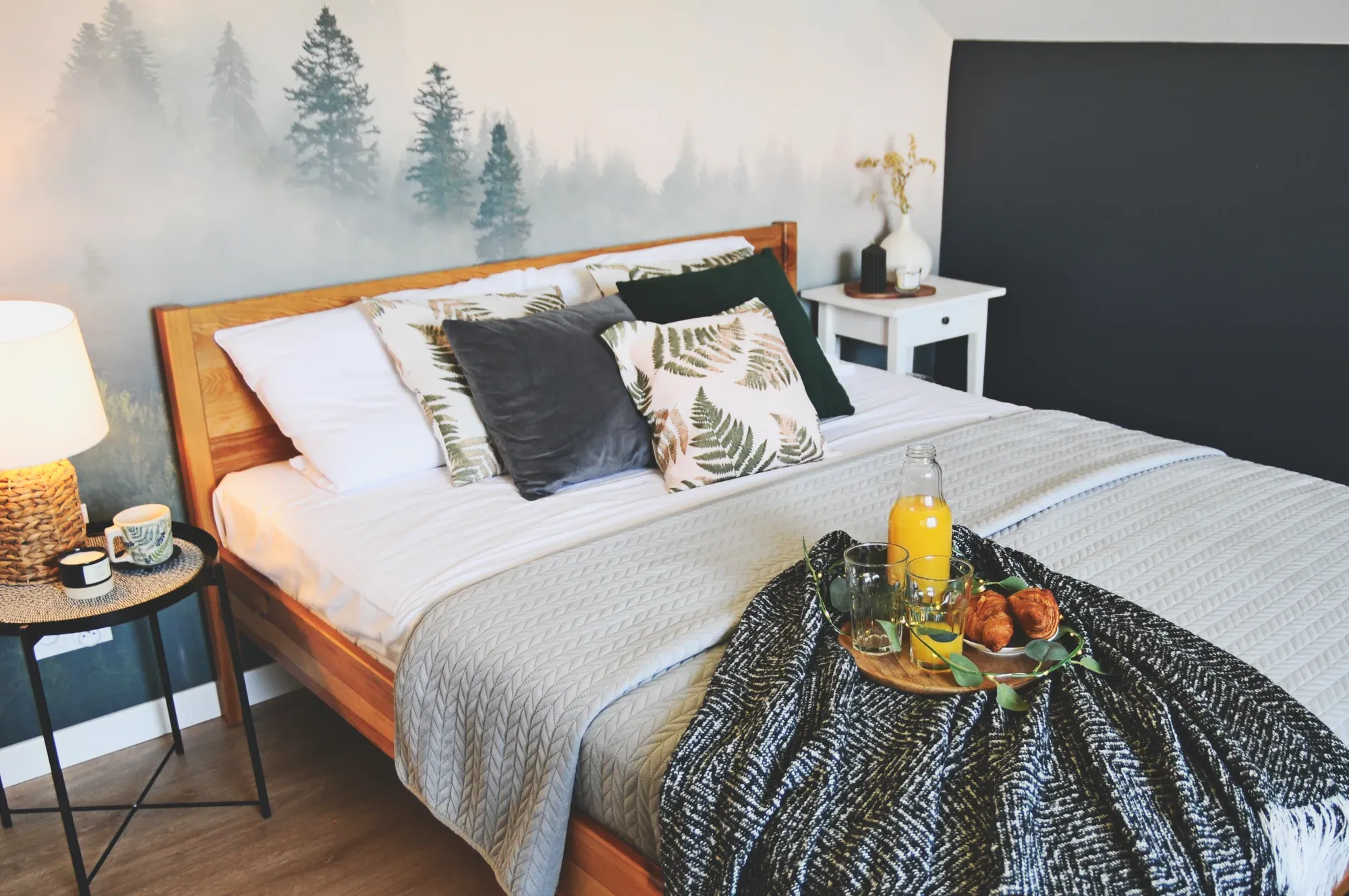 Sypialnia po home stagingu: drewniane łóżko z białą pościelą, taca ze śniadaniem, fototapeta z lasem we mgle